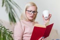 Young woman at home sitting on modern sofa relaxing in her living room reading book and drinking coffee or tea. White cozy bed and Royalty Free Stock Photo