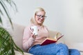 Young woman at home sitting on modern sofa relaxing in her living room reading book and drinking coffee or tea. White cozy bed and Royalty Free Stock Photo