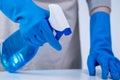 Young woman housekeeper in apron is cleaning, wiping down table surface with blue gloves, wet yellow rag, spraying bottle cleaner