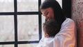 A young woman hugging with her toddler biracial son near the window