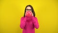 A young woman inflates a red balloon with her mouth on a yellow background. Girl in a pink turtleneck and glasses. Royalty Free Stock Photo