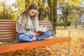 Young woman with interest reads book in autumn park. Girl sitting on bench on background of fallen yellow leaves Royalty Free Stock Photo