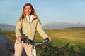 Young woman in jacket standing with a bicycle on asphalt country road, afternoon sun shines on fields meadows and mountains Royalty Free Stock Photo