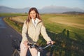 Young woman in jacket standing with a bicycle on asphalt country road, afternoon sun shines on fields meadows and mountains Royalty Free Stock Photo