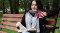 Young woman in jeans, coat and scarf, on a park bench. A woman is reading a book and drinking coffee or other hot drink outdoors Royalty Free Stock Photo