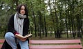 Young woman in jeans, coat and scarf, on a park bench. A woman is reading a book and drinking coffee or other hot drink outdoors Royalty Free Stock Photo