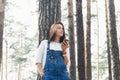 Young woman in jeans overalls with smartphone standing in woodland