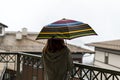 Young woman in khaki jacket from behind with rainbow umbrella standing on the terrace in the rain Royalty Free Stock Photo