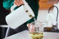 Young woman in kitchen during quarantine. Hand hold white electric smart kettle and pour hot water into teapot. Making