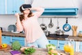 young woman with knife in virtual reality headset standing at table with fresh vegetables in kitchen Royalty Free Stock Photo