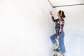 Young woman on a ladder painting new apartment walls in color white with paint roller. Renovating house