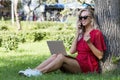 Young woman with laptop in the park. Laughing blonde with long hair in a red dress sits on the grass under a tree and talks on the Royalty Free Stock Photo