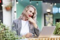 Young woman with laptop sits in a summer street cafe. Beautiful smiling blonde is running. Online training, remote work and social Royalty Free Stock Photo