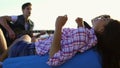 Young woman laying on easychair and listening to a man playing guitar among group of friends sitting on the beach and