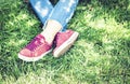 Young Woman Legs In Sport Shoes Sneakers Of Pink Suede, Sitting On The Grass Lawn In Park