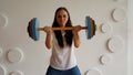 Young woman lifts multicolored barbell against white patterned wall. Adult lady doing sport, using all her strength