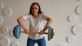 Young woman lifts multicolored barbell against white patterned wall. Adult lady doing sport, using all her strength