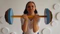 Young woman lifts multicolored barbell against white patterned wall. Adult lady doing sport, using all her strength