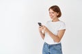 Young woman listen music in wireless headphones, looking at message on cell phone, reading screen and smiling, standing Royalty Free Stock Photo