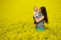 Young woman with little baby in hands in bright yellow field of flowers in sunny summer day, family love and sincerity