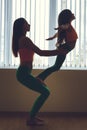 Young woman and little girl practicing hard yoga pose, silhouette near the window, full growth