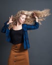 A young woman with long blond wavy hair is dancing against a dark background. Positive emotions, happy, isolated, hipster style,