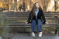 Young woman with long brown hair sits on bench in autumn park and waits for someone. Autumn time