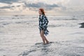 A young woman in a long checkered shirt stands on the black sand. feet in cold water, stormy waves of the sea. Heavy gray rain Royalty Free Stock Photo