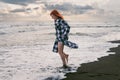 A young woman in a long checkered shirt stands on the black sand. feet in cold water, stormy waves of the sea. Heavy gray rain Royalty Free Stock Photo