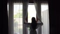 Young woman with long hair in bathrobe unveil curtains and look at the garden standing near the window in home and come