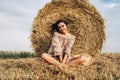 A young woman with long hair and in a dress sits near a hay bale. Woman posing smiling and looking at camera Royalty Free Stock Photo