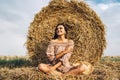 A young woman with long hair and in a dress sits near a hay bale. Woman posing smiling and looking at camera Royalty Free Stock Photo