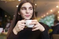 Young woman with long hair feel lonely, dreaming, drinking coffee in hands having rest in cafe near window.