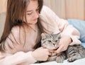 young woman with long hair lying on a bed and grey tabby cat sitting next to her. living with pet friends, relaxing Royalty Free Stock Photo