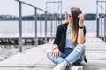 Young woman with long hair in stylish glasses posing on a wooden pier near the lake. Girl dressed in jeans and t-shirt smiling and Royalty Free Stock Photo