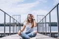 Young woman with long hair in stylish glasses posing on a wooden pier near the lake. Girl dressed in jeans and t-shirt smiling and Royalty Free Stock Photo