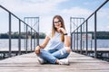 Young woman with long hair in stylish glasses posing on a wooden pier near the lake. Girl dressed in jeans and t-shirt smiling and Royalty Free Stock Photo