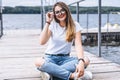 Young woman with long hair in stylish glasses posing on a wooden pier near the lake. Girl dressed in jeans and t-shirt smiling and Royalty Free Stock Photo