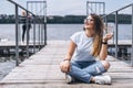 Young woman with long hair in stylish glasses posing on a wooden pier near the lake. Girl dressed in jeans and t-shirt smiling and Royalty Free Stock Photo