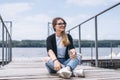 Young woman with long hair in stylish glasses posing on a wooden pier near the lake. Girl dressed in jeans and t-shirt smiling and Royalty Free Stock Photo