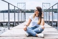 Young woman with long hair in stylish glasses posing on a wooden pier near the lake. Girl dressed in jeans and t-shirt smiling and Royalty Free Stock Photo