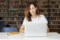 Young woman with long hair working on laptop at co-working office or library, bookshelf behind. Businesswoman reading a book