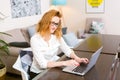 Young woman with long red hair and glasses for view works, prints on a gray laptop keyboard sitting at a wooden table in a bright Royalty Free Stock Photo