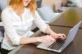 Young woman with long red hair and glasses for view works, prints on a gray laptop keyboard sitting at a wooden table in a bright Royalty Free Stock Photo