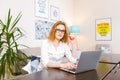 Young woman with long red hair and glasses for view works, prints on a gray laptop keyboard sitting at a wooden table in a bright Royalty Free Stock Photo