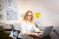 Young woman with long red hair and glasses for view works, prints on a gray laptop keyboard sitting at a wooden table in a bright Royalty Free Stock Photo