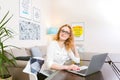 Young woman with long red hair and glasses for view works, prints on a gray laptop keyboard sitting at a wooden table in a bright Royalty Free Stock Photo