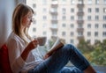 Young woman with long red hair in glasses in a white shirt sitting on the windowsill at home reading a book drinking coffee Royalty Free Stock Photo