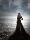 Young woman in long transparent black dress among beach and sea. Wind blows hem of her dress