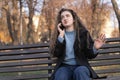 Young woman with long wavy brown hair sits on bench in park and indignaly talks on phone and gesticulating Royalty Free Stock Photo